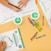 School table featuring STEM observation journals, crayons, and green lids for Cups of 5 Baby caterpillars