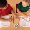 Two children at a school table with open workbooks, colored pencils, and the green top of a Cup of Caterpillars