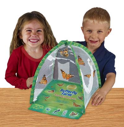 Two children smiling at camera standing behind green, tent-shaped habitat with clear viewing panel, fold-out life cycle learning tool and Painted Lady butterflies fluttering inside.