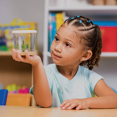 Young smiling girl watching 5 caterpillars eat their brown food in a clear cup with white lid.